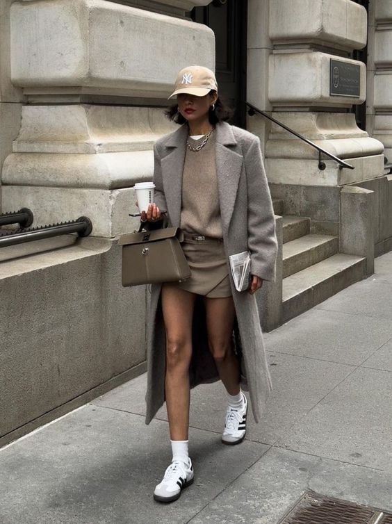 Woman in a stylish outfit with grey coat, beige shorts, and cap walking on city street holding coffee and handbag.