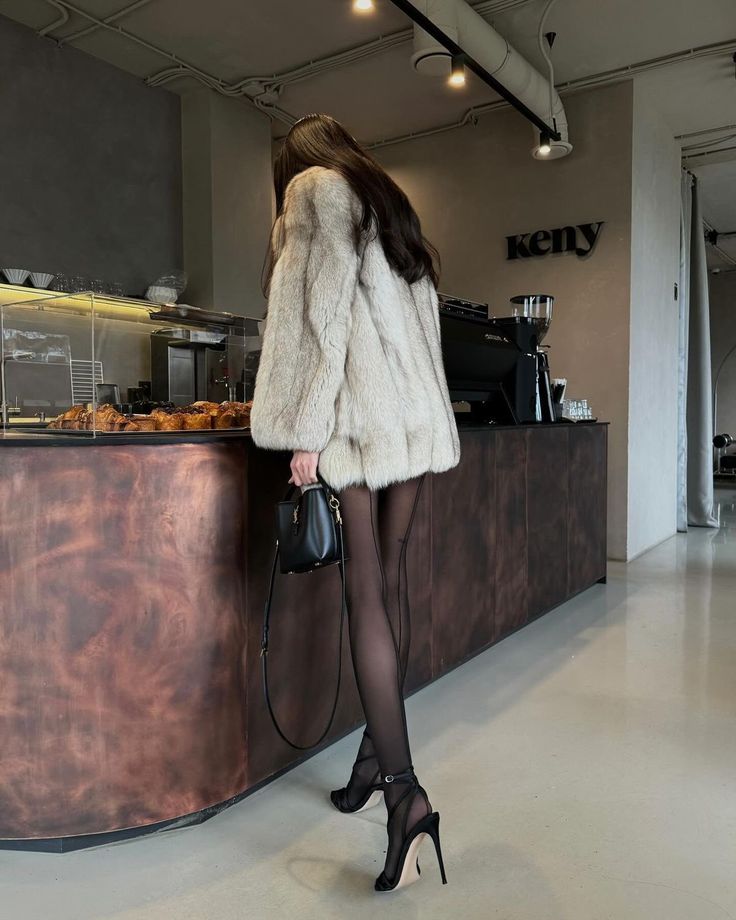 Woman in fur coat and heels standing at cafe counter with pastries and black clutch. Cozy and stylish cafe ambiance.