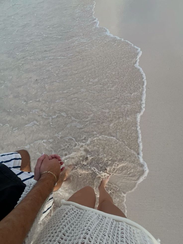 Couple holding hands and walking along the beach shore, feet in soft waves, enjoying a serene beach moment.