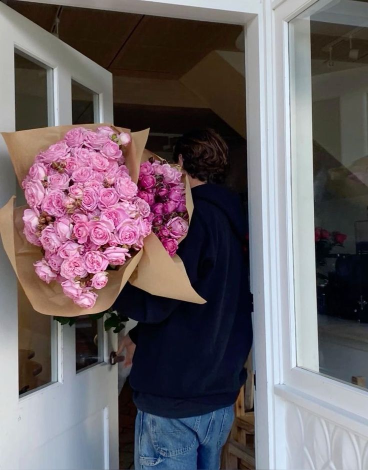 Person carrying large bouquets of pink roses through a doorway, wearing a hoodie and jeans.