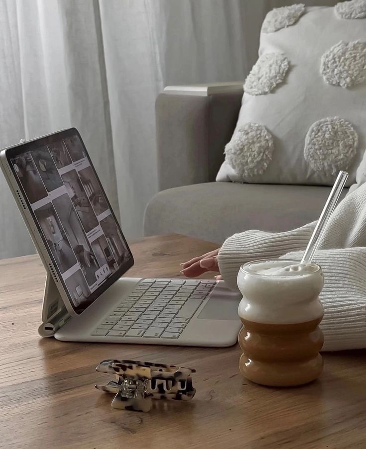 Cozy workspace with laptop, latte, and hair clip on wooden table, comfy couch in background.