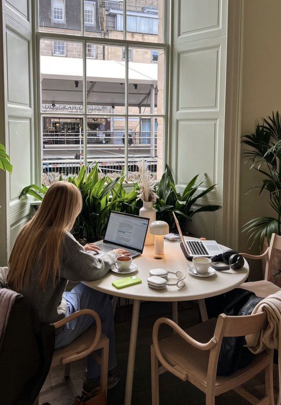 Person working on a laptop at a cozy cafe table with plants and large window in background.