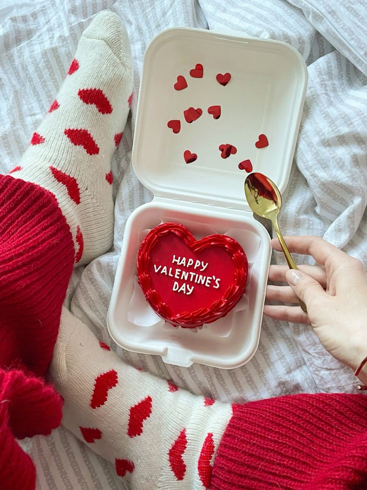 Cozy Valentine's scene with heart-shaped cake, red and white socks, golden spoon, and scattered heart confetti.