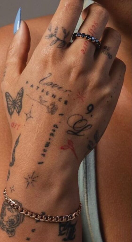 Close-up of a hand with various tattoos, including a butterfly, stars, and script, wearing rings and a chain bracelet.