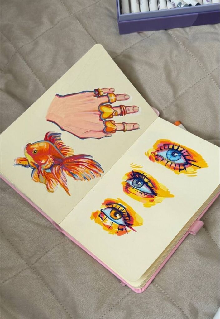 Sketchbook with vibrant drawings of a hand, fish, and eyes using colorful art pens.