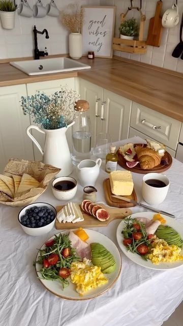 Breakfast spread in cozy kitchen with eggs, avocado, croissants, and coffee on a white tablecloth.