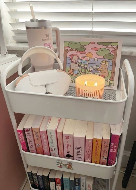 Cozy reading nook with candle, books, bag, and art on a white shelf cart by a window.