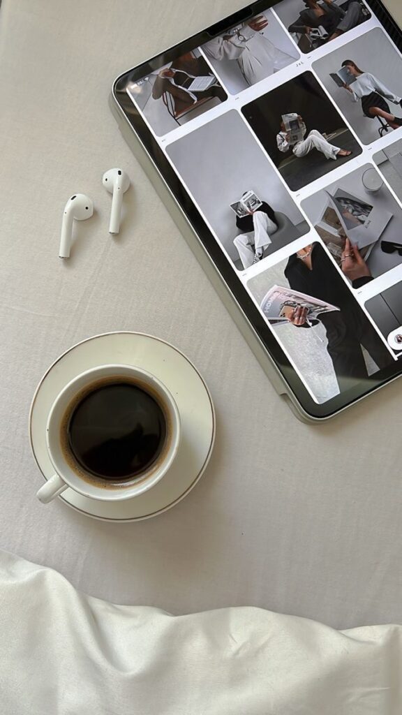 Tablet browsing fashion images with coffee and wireless earbuds on a white tablecloth.