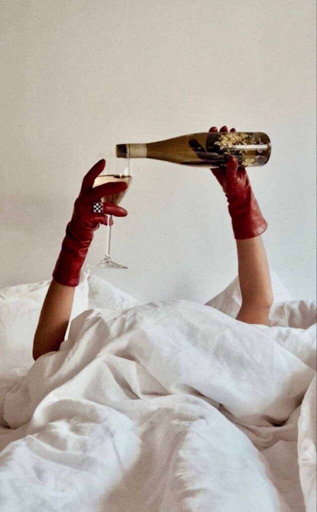 Person in red gloves pouring wine into a glass while lying in bed with white sheets.