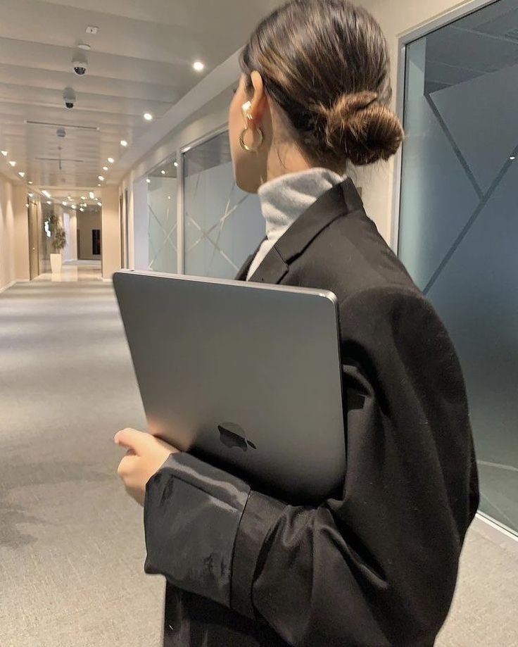 Professional woman holding a laptop in a modern office hallway, wearing a stylish black blazer and gold earrings.