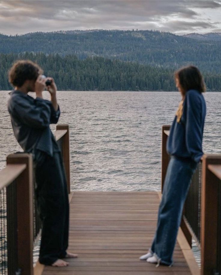 Two people on a wooden pier, with one taking a photo, overlooking a scenic lake and forested hills at dusk.