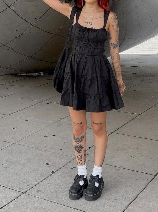 Person in a black dress with tattoos, posing near reflective sculpture, wearing black shoes and white socks.