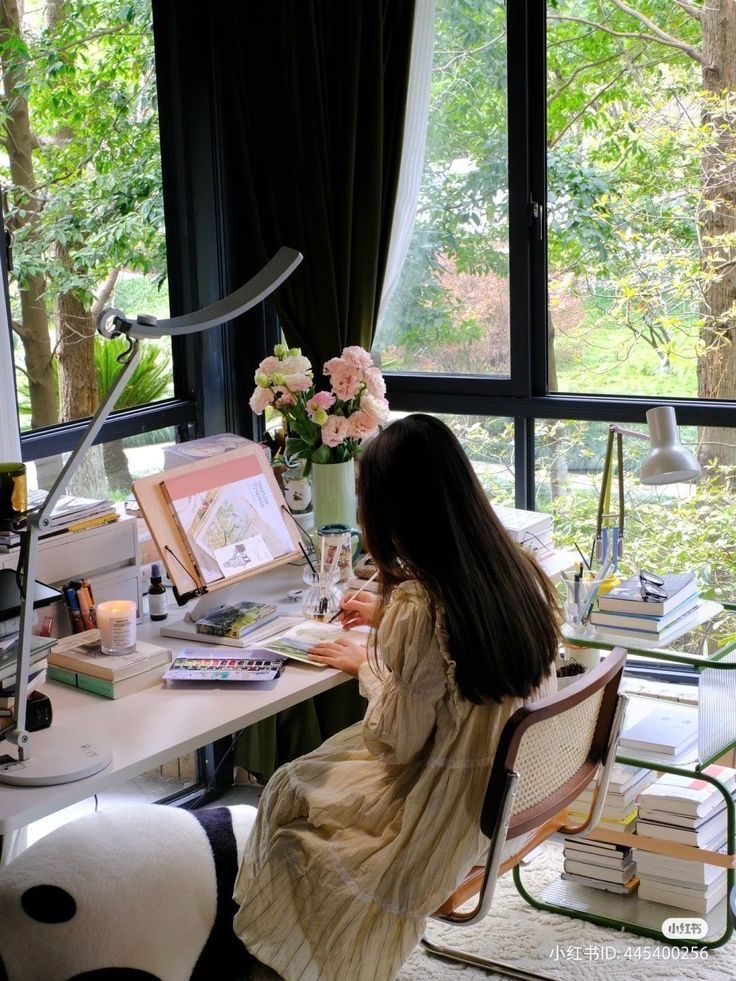 Woman painting in a bright artist's studio, surrounded by books, flowers, and nature through large windows.