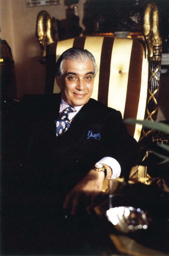 Elderly man in a suit sitting on a luxurious, striped chair with a confident smile, elegant home setting.