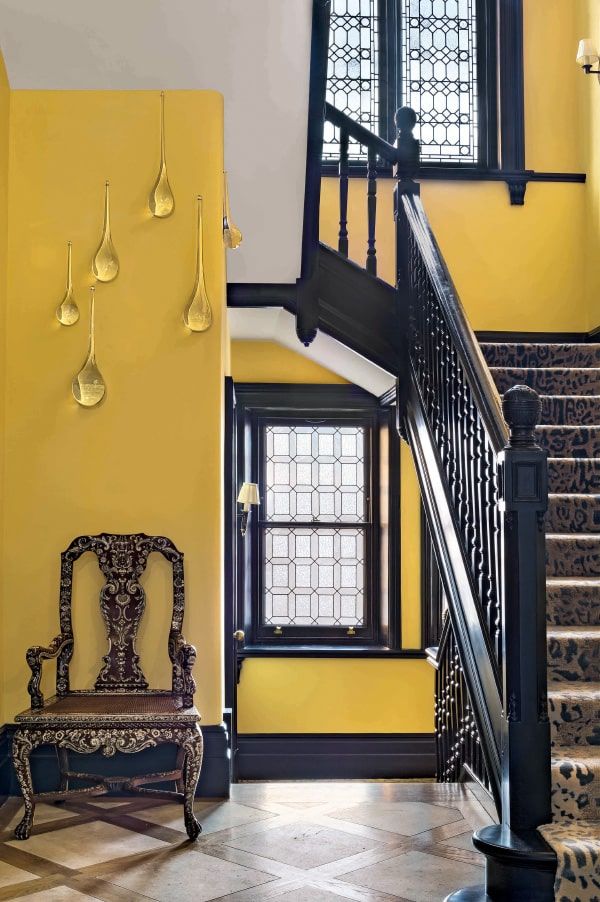 Elegant yellow hallway with ornate chair, staircase, and decorative wall art drops near window.