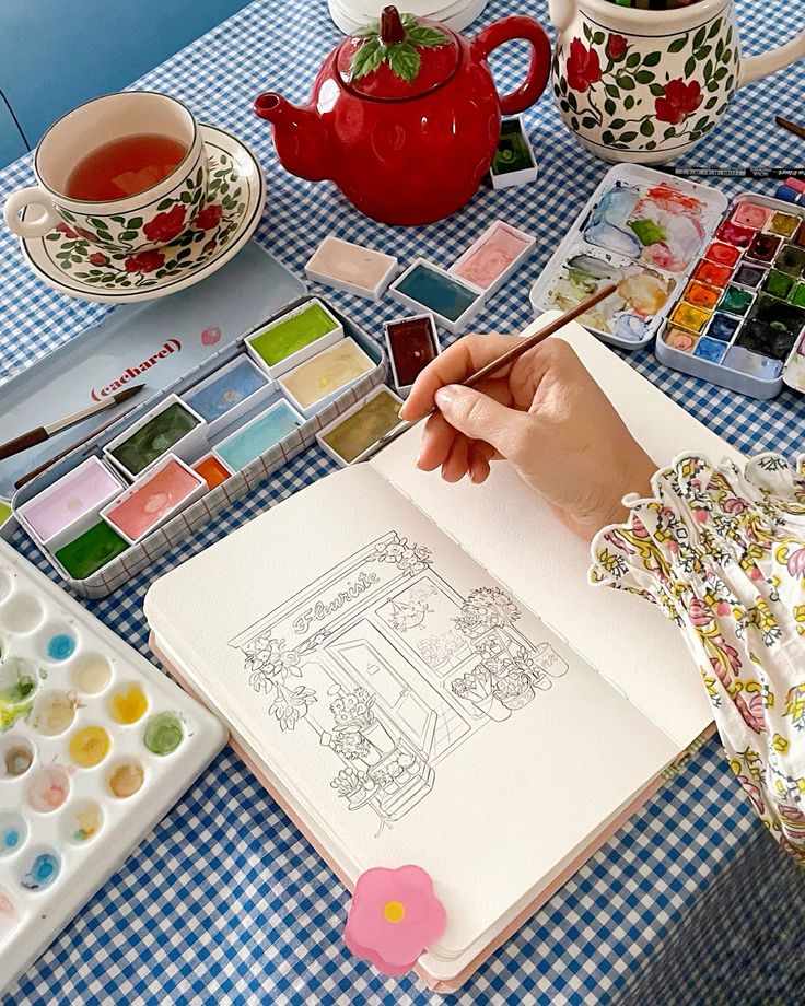 Artist painting a floral shop scene with watercolors on a gingham table, next to a red teapot and tea cup.