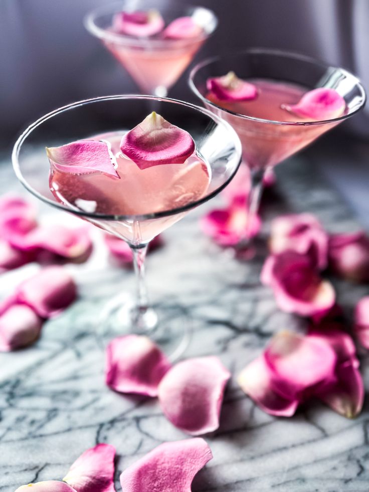 Elegant pink cocktail garnished with rose petals in martini glasses on a marble surface, surrounded by more petals.