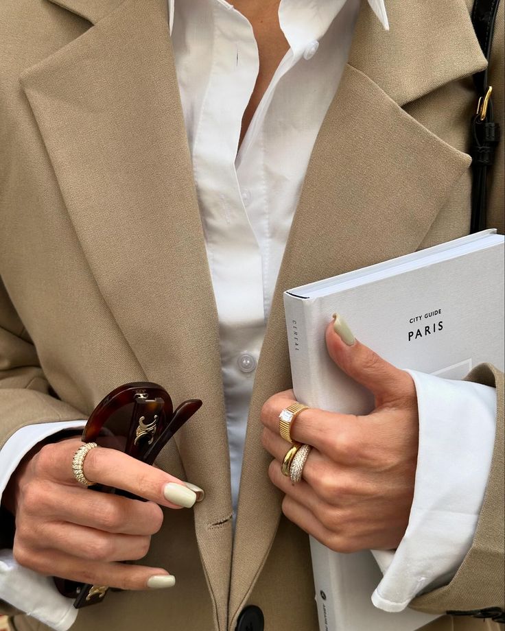 Stylish person in beige coat with sunglasses and rings holding a Paris city guidebook. Fashion-forward urban look.