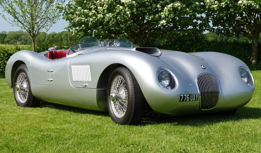 Silver vintage sports car parked on lush grass, with trees in the background under a clear sky.