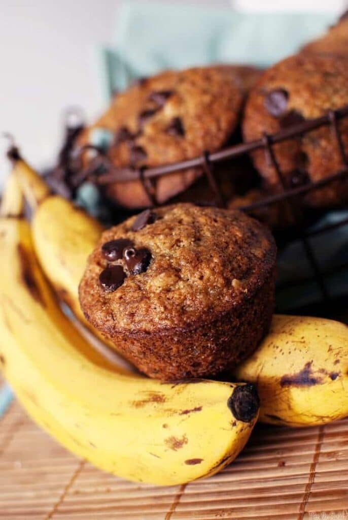 Banana chocolate chip muffins with fresh bananas in a wire basket, ideal for a tasty breakfast treat.