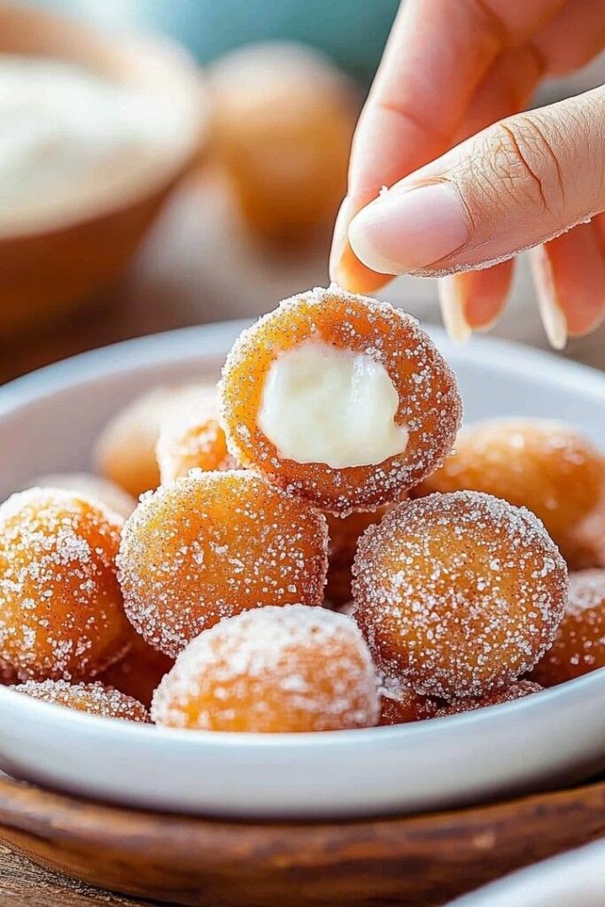 Hand holding sugar-coated doughnut hole with creamy filling on a plate. Delicious dessert treat.