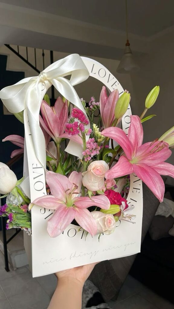 Bouquet of pink lilies and roses in a gift box with Love text, adorned with a white ribbon, indoor setting.