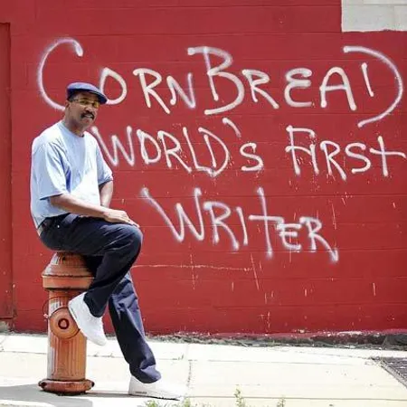 Man sitting on hydrant in front of graffiti saying Cornbread World's First Writer on a red wall.