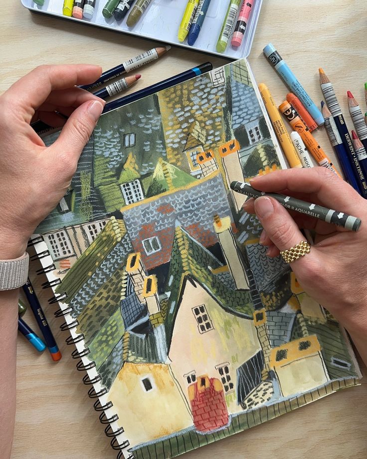 Artist sketching colorful village scene with pencils and crayons on a notepad.