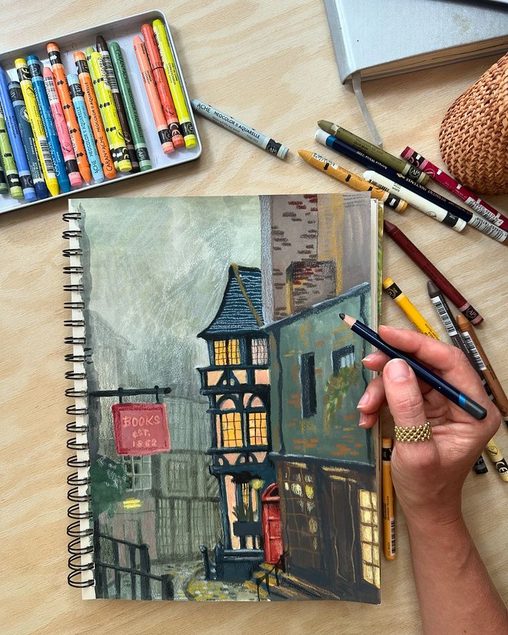 Artist drawing a quaint bookstore street scene with pastels in a sketchbook, surrounded by colorful art supplies.