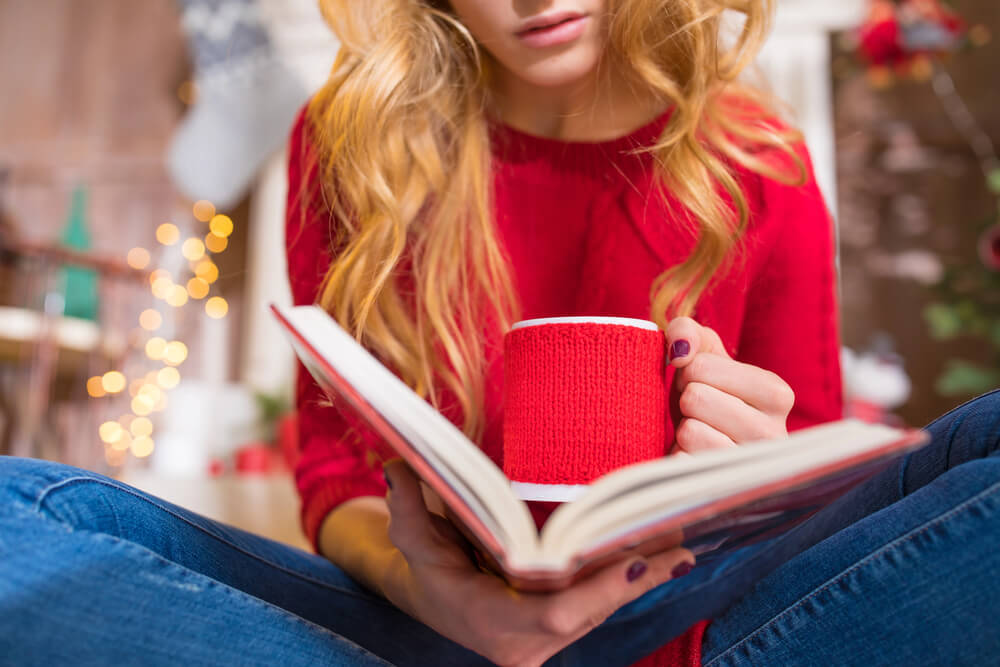 Woman in red sweater reading a book, holding a coffee mug, cozy home setting with festive lights.