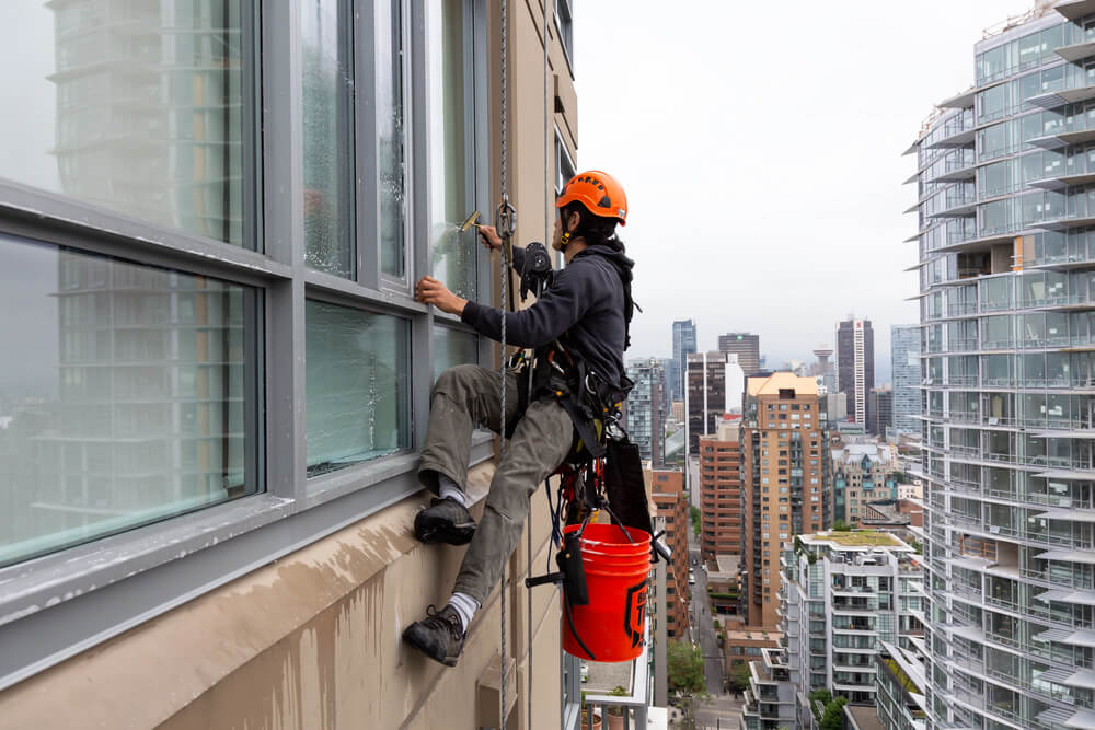 Window cleaner in safety gear working on a high-rise building with cityscape view in the background.