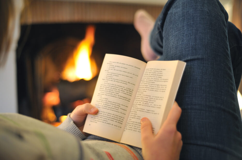 Person reading a book by a cozy fireplace, wearing blue jeans and holding an open book in their hands.