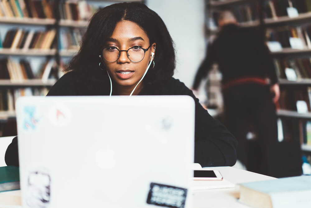 Young woman focused on laptop work in a library setting, wearing glasses and headphones.