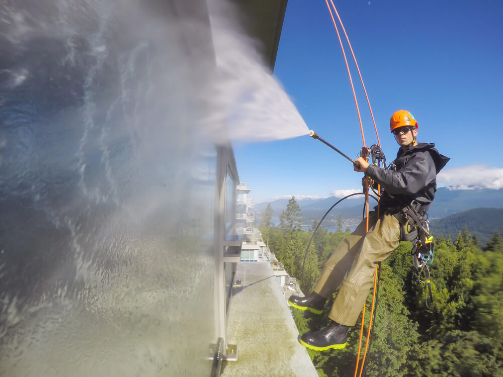 Worker on ropes pressure washing high-rise windows with scenic mountain view in the background.