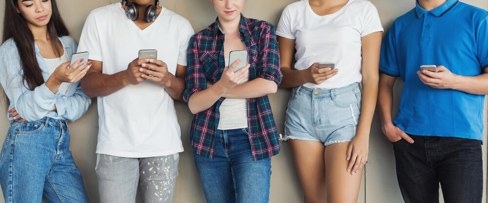 Diverse group of young people looking at smartphones, standing against a wall, casual attire.