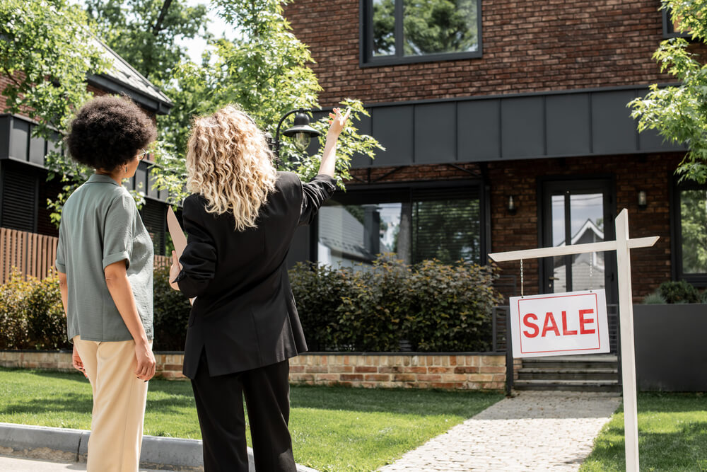 Two people view a house for sale, with a Sale sign in front, discussing potential purchase options.