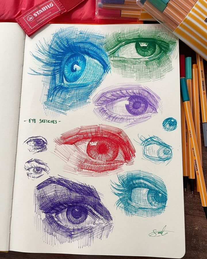 Colorful eye sketches in pen on a notebook, surrounded by art supplies, showcasing artistic techniques.