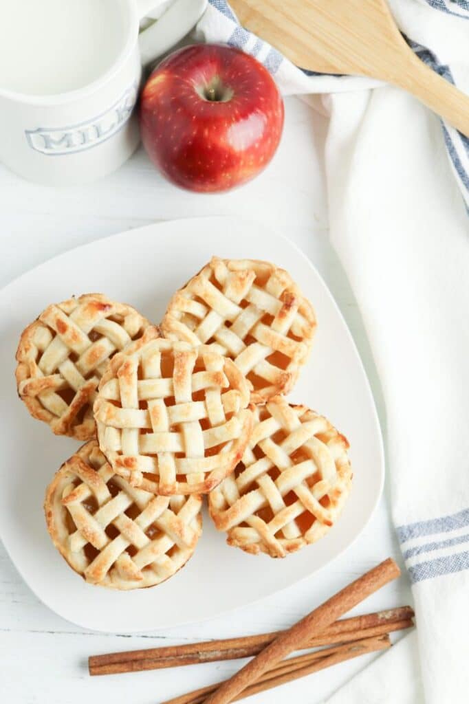 Mini apple pies with lattice crust on a white plate, surrounded by red apple, milk, and cinnamon sticks.