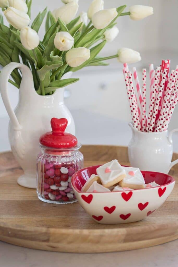 Valentine's Day setup with heart-patterned bowl, cookies, candy jar, tulips, and straws on a wooden tray.