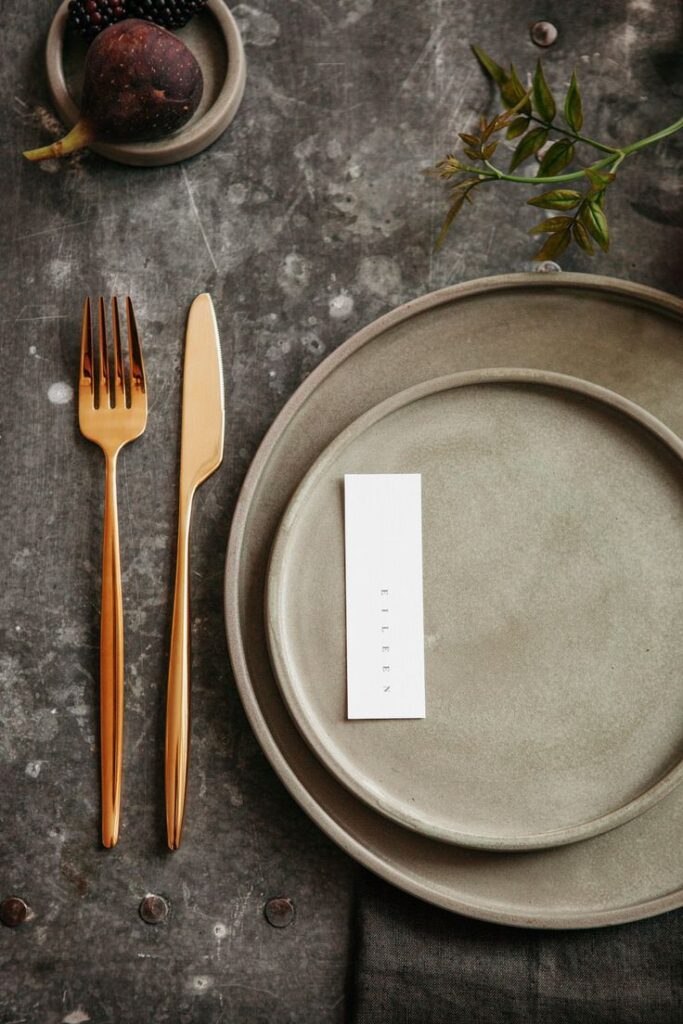 Elegant table setting with rustic plates, gold cutlery, fig, and a name card on a textured surface.