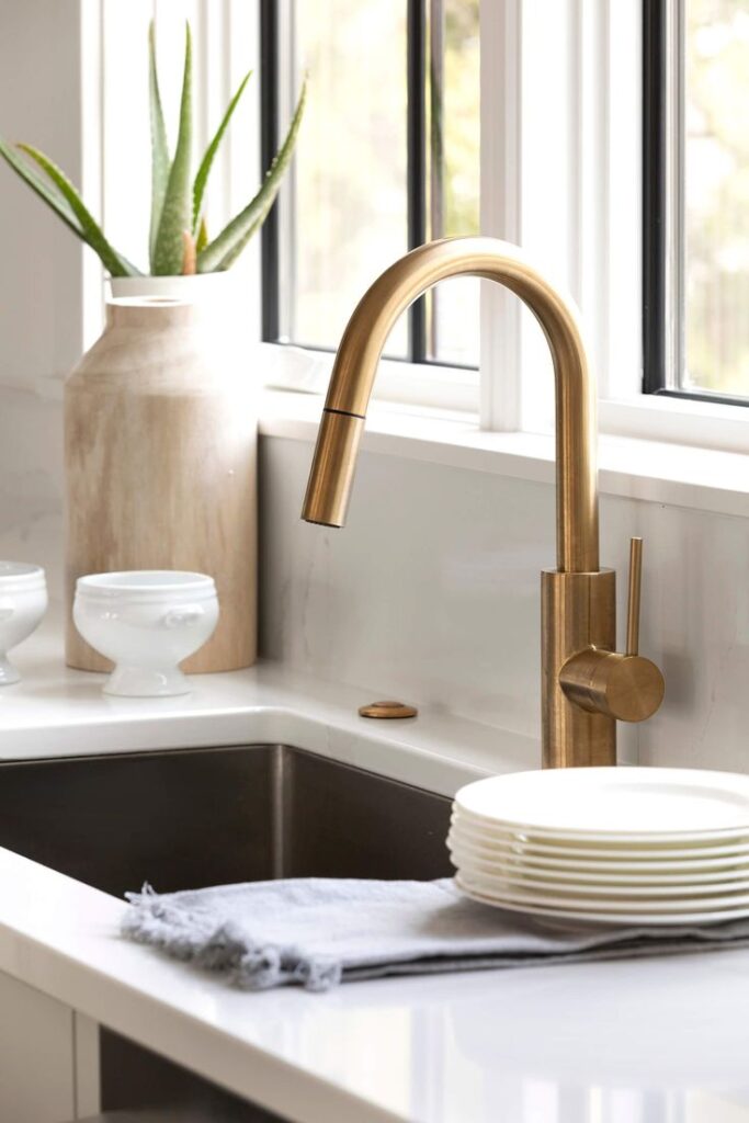 Brass kitchen faucet with white sink, plates, aloe plant, and large window in bright, modern kitchen.