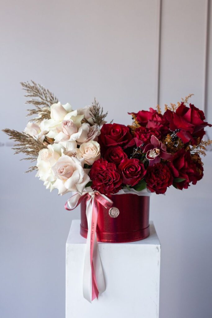 Elegant floral arrangement with red and white roses in a decorative red box on white pedestal.