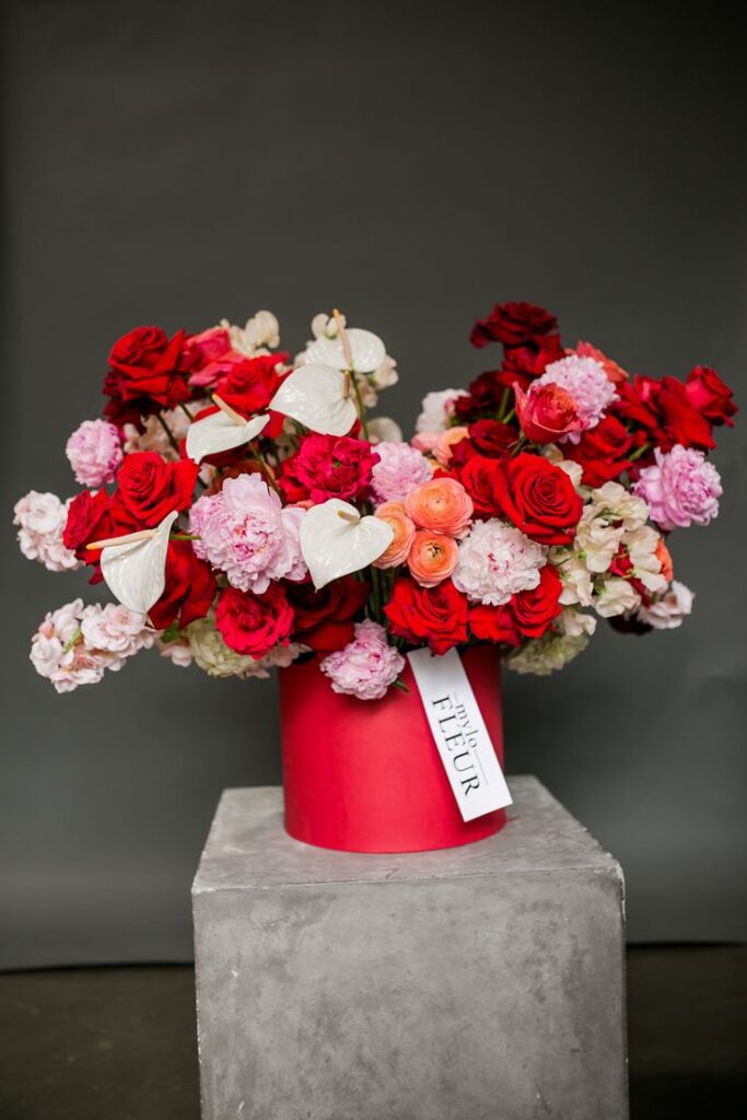 Elegant floral arrangement with red roses and pink peonies in a red vase on a gray platform.