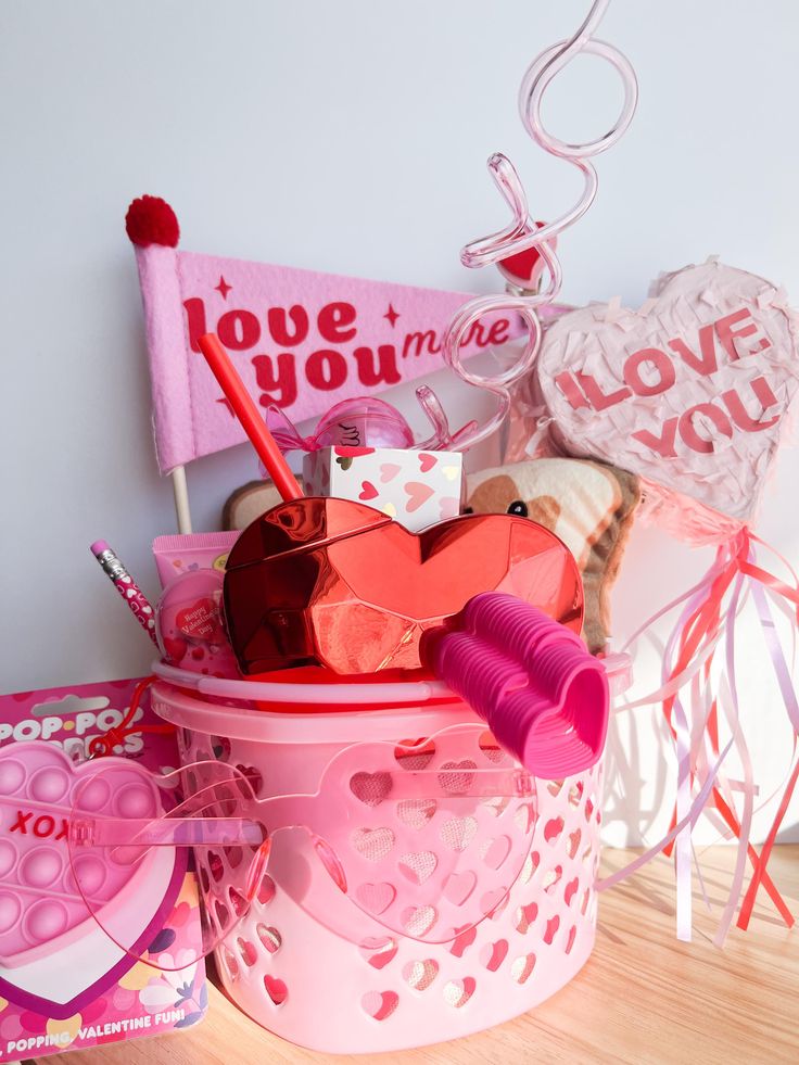 Pink Valentine's Day gift basket with heart-themed items, including glasses, a flag, and candy, on a wooden table.