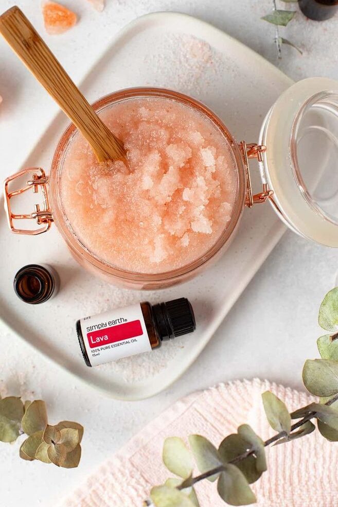 Jar of pink Himalayan salt scrub with wooden spoon, surrounded by essential oil bottle and eucalyptus leaves.