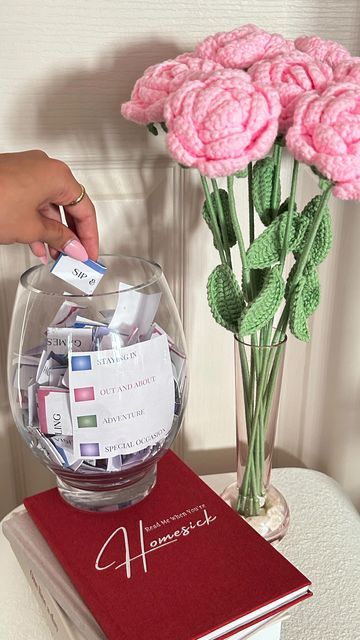 Glass jar with activity slips, pink crochet flowers in a vase, and a red journal on a table. Cozy creative setup.