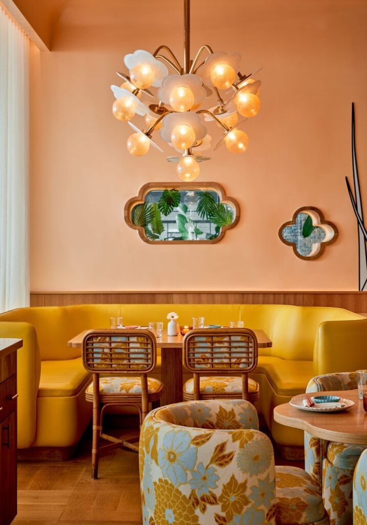 Chic restaurant interior with yellow seating, retro chandelier, floral chairs, and unique wall mirrors.