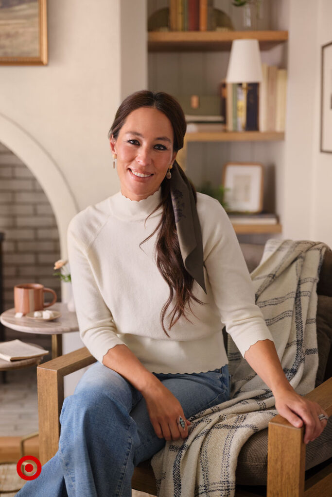 Smiling woman in casual attire sitting in a cozy living room with bookshelves and a fireplace.