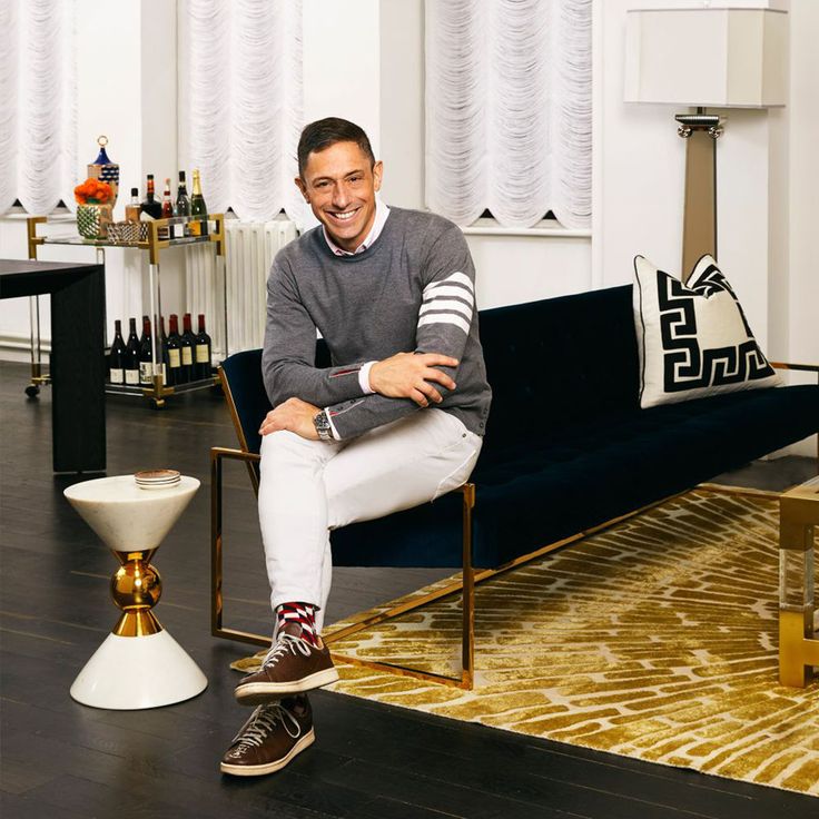 Man in stylish modern living room, sitting on dark sofa, wearing gray sweater and white pants, with decorative elements.