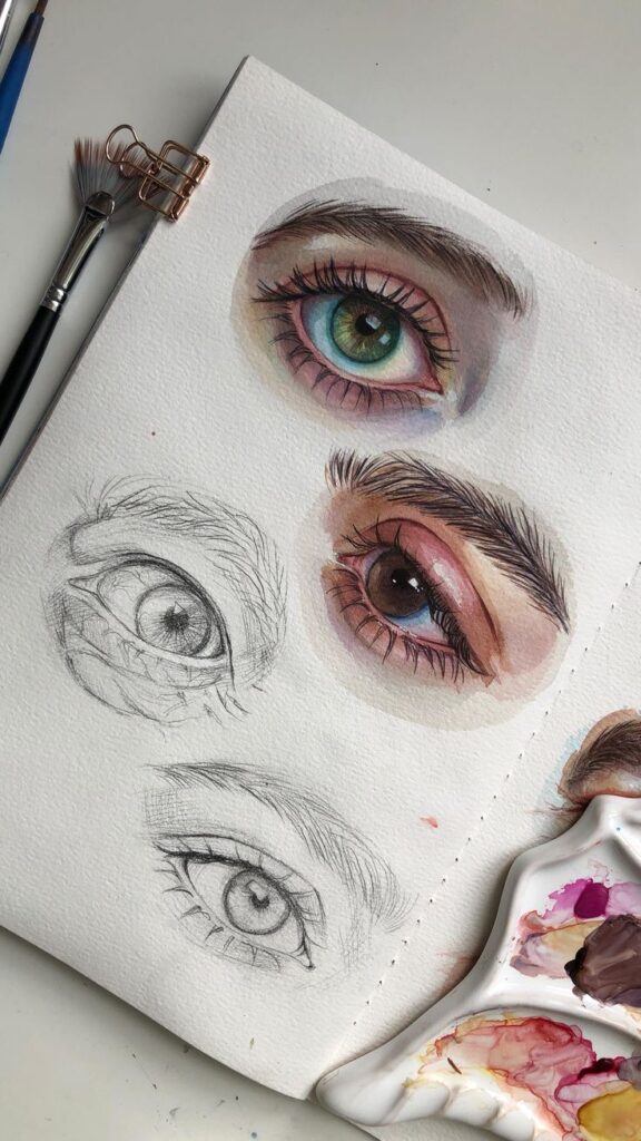 Watercolor and pencil eye sketches on paper with paintbrushes and palette. Sketchbook art in progress.